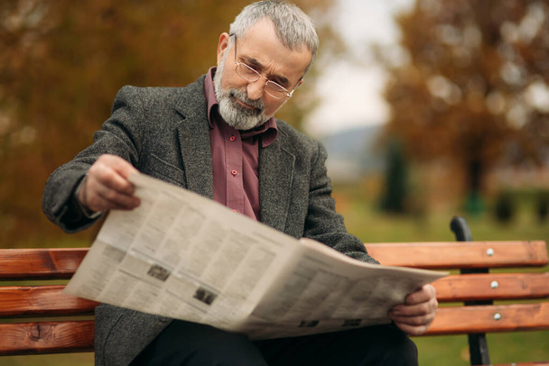 Un gentil grand-père avec une belle barbe dans une veste grise s'assoit sur un banc dans le parc et lit un journal
. - Photo, image