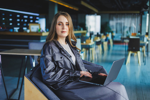 Uma jovem mulher em uma jaqueta de couro senta-se em um café e trabalha em um laptop. Menina bonita em roupas casuais. - Foto, Imagem