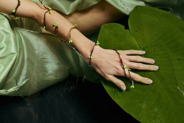 A womans hand adorned with a delicate green bracelet rests on a large lily pad. - Photo, Image