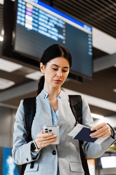 Atrakcyjna kobieta stojąca z biletem i paszportem na lotnisku. Azjatka w tle rozkładu lotów. Turysta czeka na rozpoczęcie 2024 letnich wakacji - Zdjęcie, obraz
