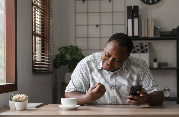 Афроамериканец сидит за столом в домашнем офисе, смотрит на кредитку, держа в руках смартфон и используя ноутбук, яркий оконный свет - Фото, изображение