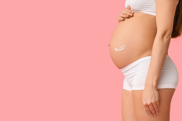 Jonge zwangere vrouw met crème tegen striae op haar buik tegen roze achtergrond - Foto, afbeelding