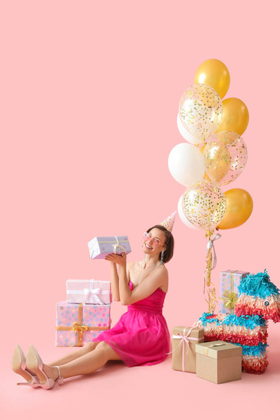 Hermosa mujer en sombrero de fiesta con regalos de cumpleaños y globos sobre fondo rosa - Foto, imagen