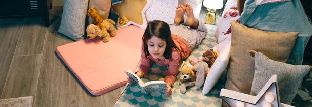 Sztandar spokojnej dziewczynki patrząc na książkę podczas odpoczynku otoczony poduszkami w tipi. Czytanie dla dzieci i relaks leżąc nad wycieraczką w przytulnym namiocie. Koncepcja pobytów i wakacji domowych. - Zdjęcie, obraz