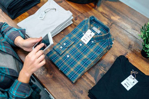 Unrecognizable female entrepreneur checking merchandise stock on her phone next to apparel stacks over counter on clothes store. Woman taking photo of plaid shirt in local shop. Small business concept - Photo, Image