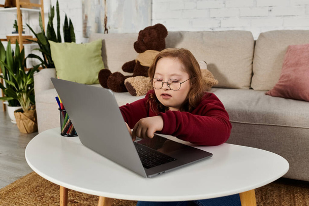 A little girl with Down syndrome uses a laptop in her living room. - Foto, afbeelding