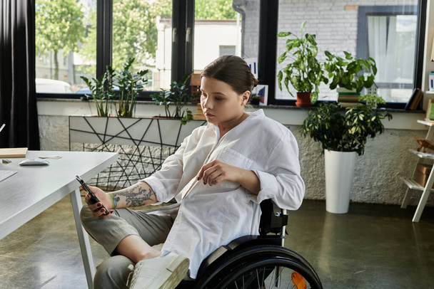 A young businesswoman in a wheelchair uses her phone while sitting at a modern desk in a bright office setting. - Foto, imagen