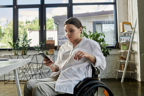 Μια νεαρή επιχειρηματίας με αναπηρία κάθεται σε αναπηρικό καροτσάκι, δουλεύοντας σε ένα σύγχρονο γραφείο. - Φωτογραφία, εικόνα