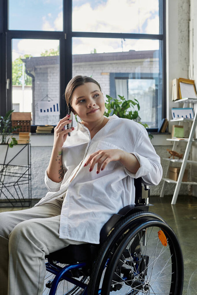 Eine junge Geschäftsfrau im Rollstuhl telefoniert während ihrer Arbeit in einem modernen Büro mit ihrem Handy. - Foto, Bild