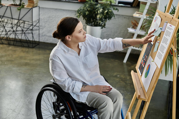 Молодая деловая женщина в инвалидной коляске работает за своим столом в современном офисе. Она сосредоточена на своей работе и кажется уверенной и способной. - Фото, изображение