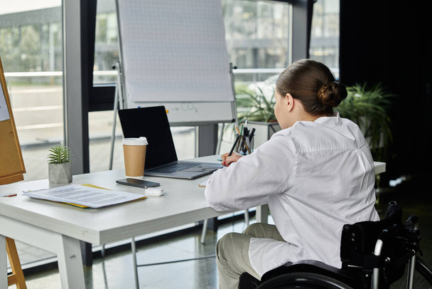 Μια νεαρή επιχειρηματίας με αναπηρία κάθεται σε αναπηρικό καροτσάκι, δουλεύοντας σε ένα γραφείο σε ένα σύγχρονο γραφείο. - Φωτογραφία, εικόνα