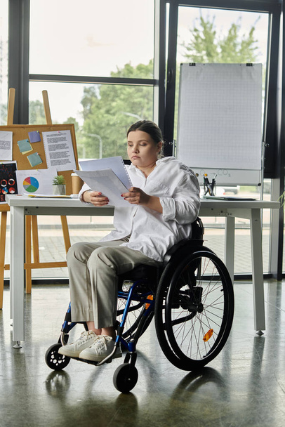 Pyörätuolissa istuva nuori liikenainen istuu työpöydän ääressä modernissa toimistossa tarkistamassa asiakirjoja.. - Valokuva, kuva