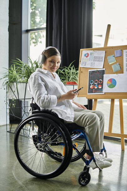 Młoda bizneswoman w słuchawkach siedząca na wózku inwalidzkim pracuje w nowoczesnym biurze. - Zdjęcie, obraz