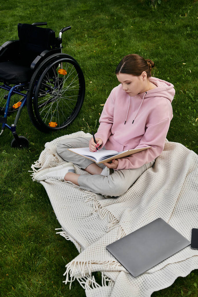 Une jeune femme avec un sweat à capuche rose est assise dans un fauteuil roulant sur une couverture dans un parc herbeux, écrivant dans un carnet. - Photo, image