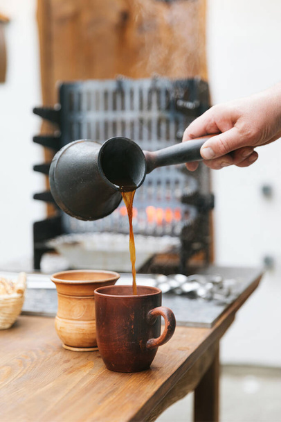 A hand pours hot coffee from a jezvah into a clay mug. A jezvah against the background of a brazier, in which fire and smoke are visible. Cope space. Vertical photo.  - Photo, Image