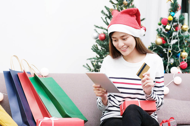 Mladý roztomilý asijské žena nosí vánoční klobouk s úsměvem při držení tablet a kreditní karty, vánoční nákupy on-line koncept - Fotografie, Obrázek