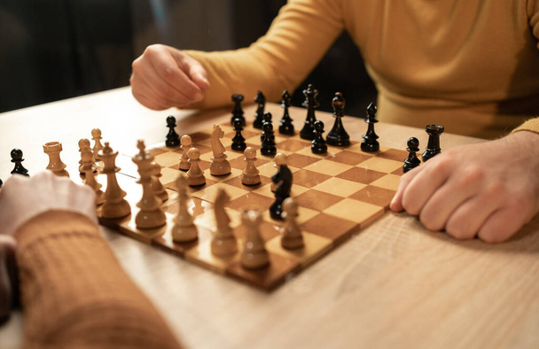 Крупним планом двоє чоловіків грають в шахи вдома. Основна увага приділяється їх рукам і дерев'яній шахівниці, коли вони роблять стратегічні ходи в приміщенні. Концепція хобі та дозвілля - Фото, зображення