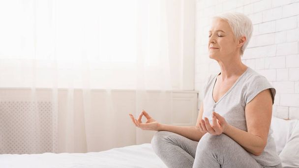 Senior vrouw doet yoga, mediteren in de lotuspositie op bed thuis, lege ruimte - Foto, afbeelding