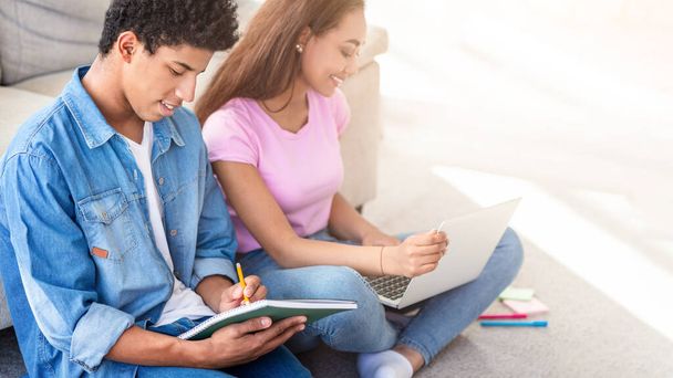 Czarna para nastolatków siedzi w domu na podłodze, skupiona na nauce. Facet pisze w notatniku, podczas gdy dziewczyna używa laptopa.. - Zdjęcie, obraz