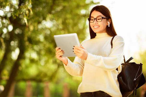 Aziatische jonge vrouw met een bril en draadloze oordopjes gebruikt een tablet terwijl ze in een zonnig park staat. Ze heeft een rugzak en lacht, geniet van de buitenomgeving. - Foto, afbeelding