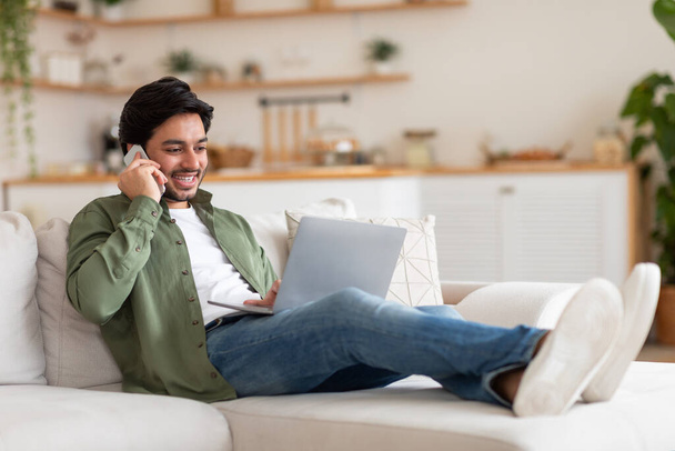 Fiatalember beszél telefonon, dolgozik Laptop a nappaliban, fekvő kanapén otthon, másolás tér - Fotó, kép