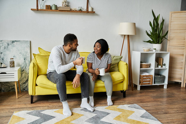 Ένα ερωτευμένο ζευγάρι Αφροαμερικανών κάθεται σε έναν κίτρινο καναπέ, απολαμβάνοντας καφέ μαζί στο σπίτι τους. - Φωτογραφία, εικόνα