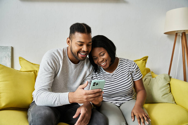 Ένα ζευγάρι κάθεται σε ένα κίτρινο καναπέ, γελώντας κοιτάζοντας ένα τηλέφωνο. - Φωτογραφία, εικόνα