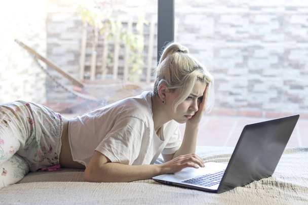 Blondine vrouw met een paardenstaart, in een wit t-shirt en bloemenpyjama, liggend op een bed, werkend aan een laptop. Ze lijkt bezorgd, met een bedachtzame uitdrukking. - Foto, afbeelding