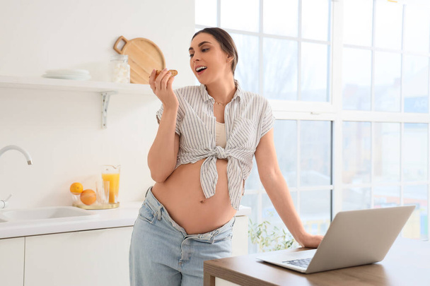 Młoda kobieta w ciąży z tostem za pomocą laptopa w kuchni - Zdjęcie, obraz