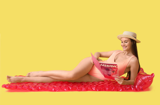 黄色の背景に対して膨脹可能なマットレスに横たわる帽子と雑誌で水着の若い女性を笑う - 写真・画像