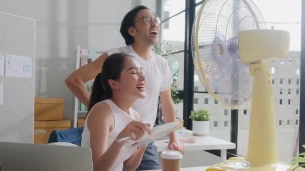 Dva mladí asijští startup spolupracovníci jsou zábavné foukání vítr s elektrickým ventilátorem, pracuje v horkých teplotách v příležitostné malé obchodní kanceláře v létě, osvěžující z nepříjemného teplého počasí. - Záběry, video