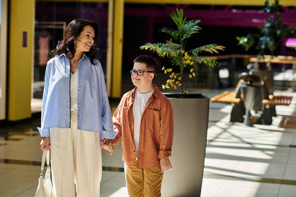 Мать и ее сын с синдромом Дауна идут рука об руку через торговый центр. - Фото, изображение