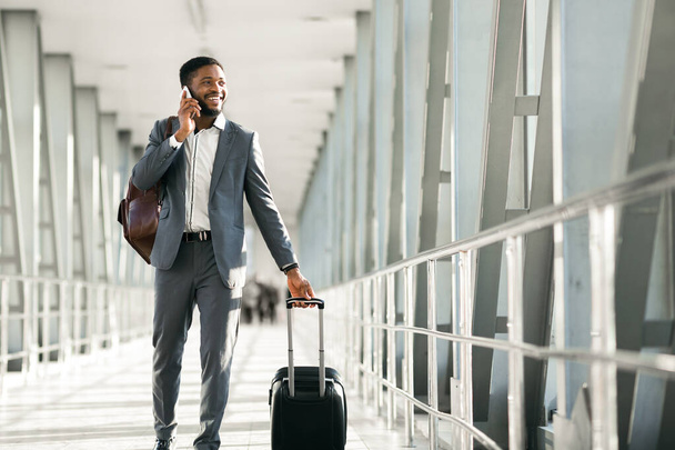 Afro-Amerikaanse man die belt met zijn bagage op een modern vliegveld. De stemming is druk en gericht, het vastleggen van de essentie van professionele reizen en communicatie. - Foto, afbeelding