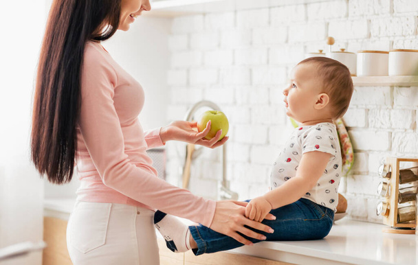 Uma mulher está segurando suavemente uma maçã em direção a um bebê, que está estendendo a mão para tocar a fruta. O bebê aparece curioso e animado, enquanto a mulher parece carinhosa e carinhosa.. - Foto, Imagem