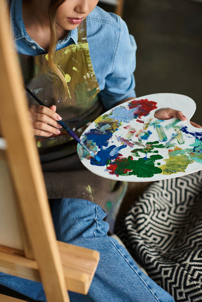 donna ritagliata in un grembiule si siede in uno studio, mescolando i colori sulla sua tavolozza. - Foto, immagini