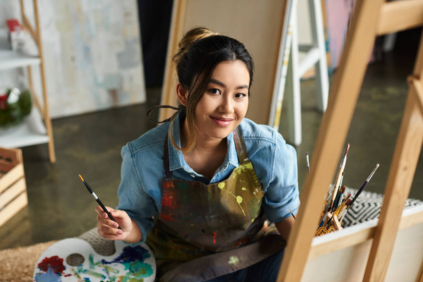 Una joven artista asiática sonríe sosteniendo un pincel y de pie frente a un caballete en su estudio de arte. - Foto, imagen