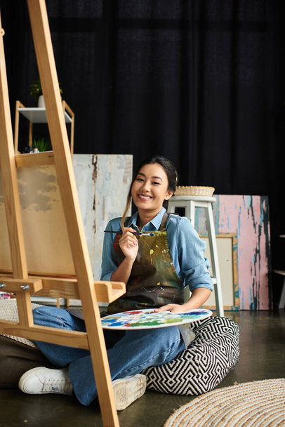 Une jeune femme asiatique dans un tablier s'assoit par terre dans son atelier d'art, tenant un pinceau et une palette, souriant à la caméra. - Photo, image
