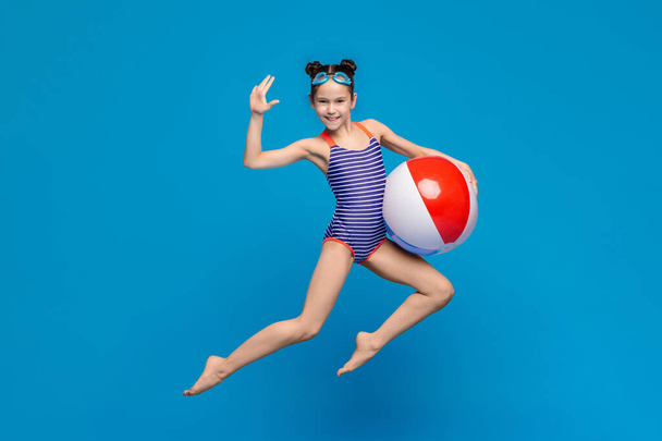 Ein junges Mädchen in einem bunten Badeanzug steht am Strand und hält freudig einen Strandball in der Hand. Der pulsierende Beachball kontrastiert mit blauem Himmel und sandigem Ufer. - Foto, Bild