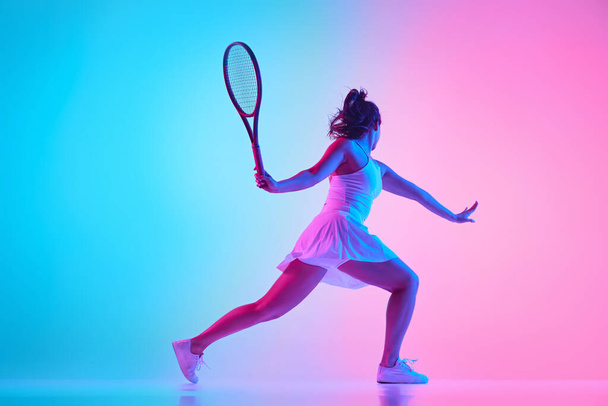 Side view tenisz sportoló fehér ruhában lengeti az ütőt, hogy erős lövés neon fényben élénk gradiens stúdió háttér. Fogalom a profi sport, bajnokság, bajnokság. - Fotó, kép