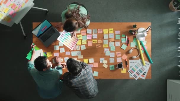 Top down view of business team with casual outfit looking laptop screen and sharing idea. Aerial view of manager working and checking color palette at table with sticky notes and headphone. Symposium. - Footage, Video