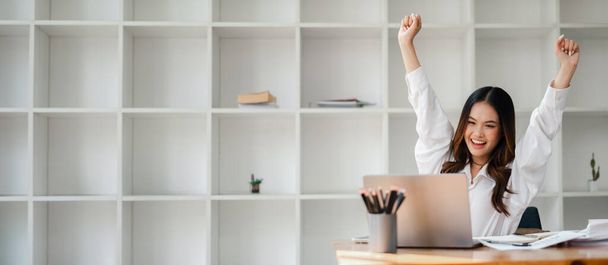 Jovem feliz levantando braços em celebração enquanto trabalhava em um laptop em um escritório moderno com prateleiras brancas. - Foto, Imagem