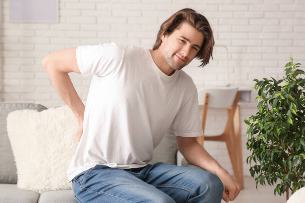 Handsome young man sitting on sofa and suffering from back pain at home - Fotografie, Obrázek