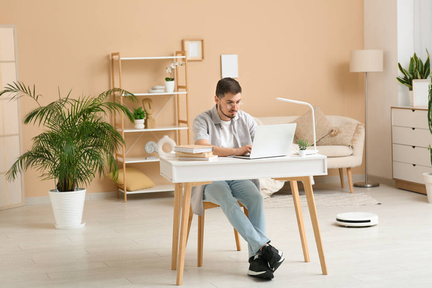 Young man using laptop at table with modern robot vacuum cleaner on floor in home office - Foto, Imagen