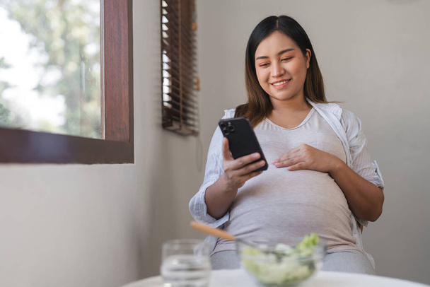 Těhotná žena sedí pohodlně v moderním obývacím pokoji, používá svůj smartphone a těší zdravé svačinu. Přírodní světlo vyplňuje útulné domácí prostředí. - Fotografie, Obrázek