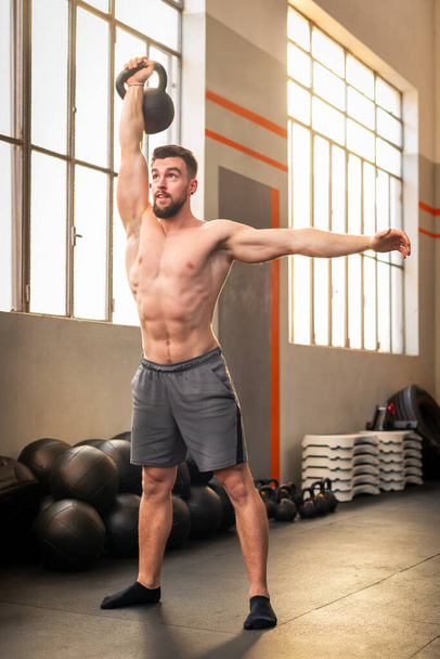 Πλήρες σώμα shirtless μυώδης γενειοφόρος ενήλικας αθλητής κοιτάζοντας μακριά, ενώ στέκεται στο πάτωμα του γυμναστηρίου κάνει ένα χέρι kettlebell αρπαγή γυμναστική στο φως της ημέρας - Φωτογραφία, εικόνα