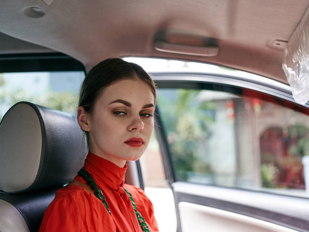 Une femme sophistiquée dans une superbe robe rouge ferme les yeux, perdue dans la pensée alors qu'elle est assise sur le siège arrière d'une voiture de luxe. - Photo, image