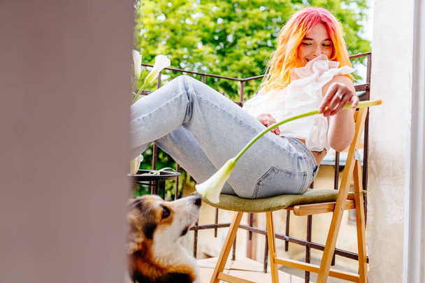 Mulher brincalhona com cabelo rosa está sentada em uma cadeira com uma flor na mão. Um cão está atrás dela, a olhar para ela. A cena é pacífica e relaxante, feminina desfrutando do tempo ao ar livre - Foto, Imagem