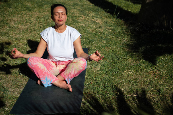 Kobieta ćwicząca jogę w pozycji siedzącej na macie do jogi w słonecznym otoczeniu na świeżym powietrzu, promująca relaks i uważność. - Zdjęcie, obraz