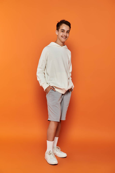 Een jonge jongen in een witte hoodie staat tegen een oranje achtergrond. - Foto, afbeelding