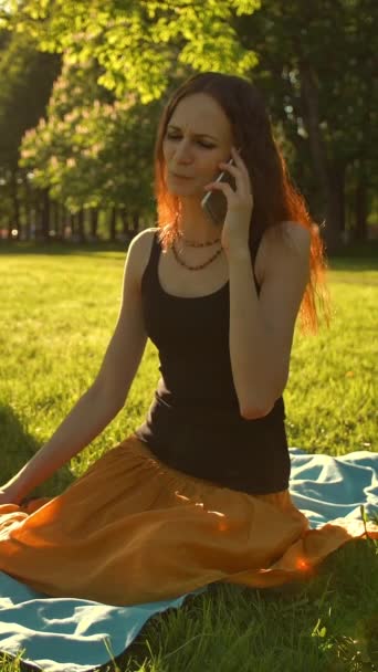 Вид спереди женщины, сидящей перед камерой и говорящей во время автофона, звонящей своей подруге в парке. - Кадры, видео